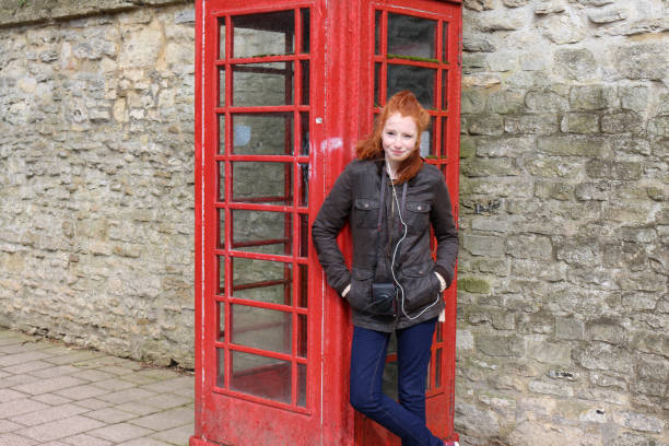Stock photo showing a beautiful, ginger haired teenager modelling ponytail hairstyle whilst stood beside a decommissioned red telephone box on an English street. The model is wearing a green wax jacket over blue denim jeans with tartan canvas shoes and a camera case slung around her neck. imagen de adolescente pelirroja adolescente 14 / 15 de pie junto a una caja de teléfono rojo desmantelado en una calle inglesa, usando una chaqueta de cera verde sobre jeans de mezclilla azul con zapatos de lona tartán y una caja de cámara colgada alred
