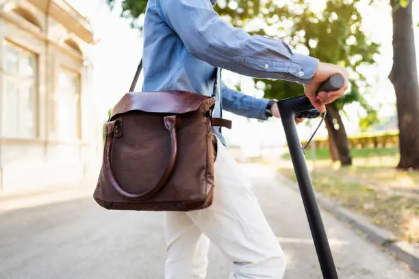 Close-up shot of an unrecognizable young man carrying a shoulder leather bag and riding an electric push scooter.