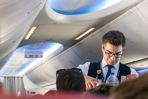 Istambul, Turkey - July 27, 2019: A young male flight attendant serving drinks to a passenger onboard a economy class cabin.