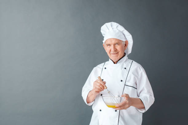 istock Senior chef studio standing isolated on gray holding whisk mixing eggs looking camera playful 1171824739