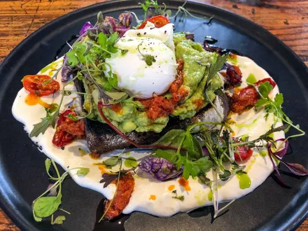 Popular brunch food: smashed avocado on dark rye toast with a poached egg, mixed salad greens, sundried tomato, olive oil, labneh
