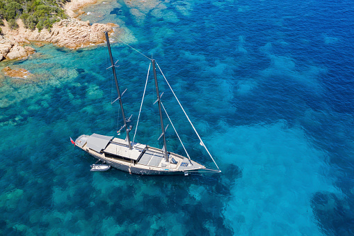 View from above, stunning aerial view of a luxury sailboat floating on a beautiful turquoise clear sea that bathes the green and rocky coasts of Sardinia. Emerald Coast (Sardinia) Italy.