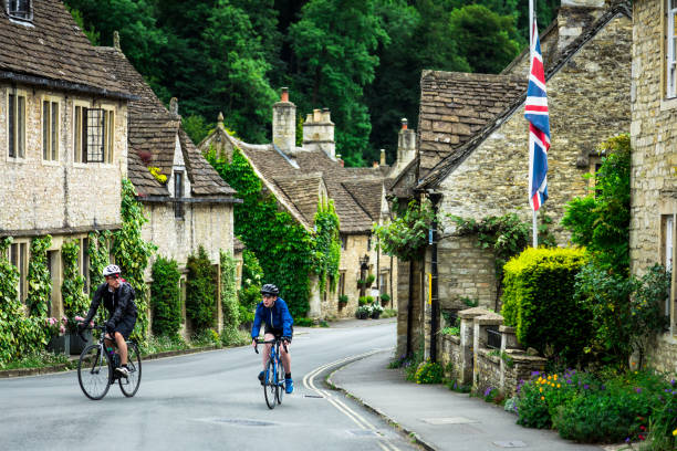 ciclisti che attraversano il tradizionale villaggio inglese con accoglienti cottage - cotswold foto e immagini stock