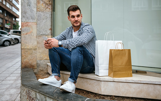 Young man with shopping bags sitting on the curb of a shop window