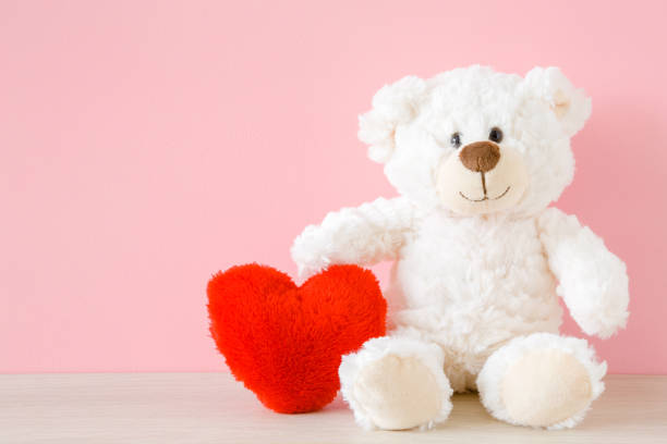 istock Smiling white teddy bear sitting and holding red soft heart at pastel pink wall. Front view. Closeup. 1171876089
