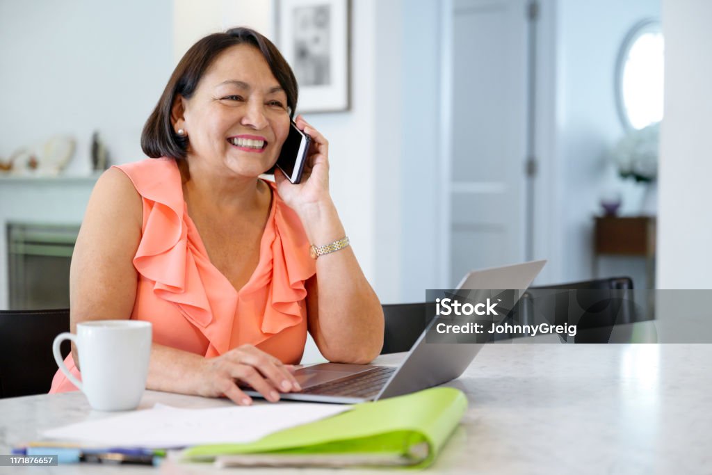 Latin American senior woman hearing good news on smart phone Hispanic senior woman using laptop at dining table and smiling as she hears good news on smart phone. Using Phone Stock Photo