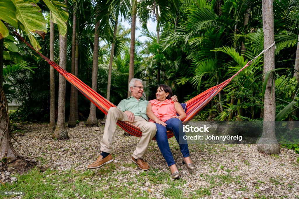 Smiling Latin American seniors reclining in backyard hammock Hispanic senior male and female relaxing in a hammock tied between trees in lush Miami backyard. Florida - US State Stock Photo
