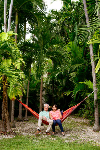 Relaxed Latin American seniors looking at camera while swinging in hammock tied between trees in Miami backyard.