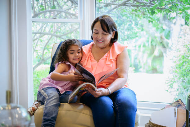 Hispanic grandmother and granddaughter enjoying picture book Young Latin American granddaughter sharing a chair with her grandmother and looking at a picture book together. candid bonding connection togetherness stock pictures, royalty-free photos & images