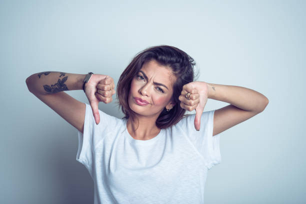 Portrait of disappointed young woman standing with thumbs down Studio portrait of displeased young asian woman wearing white t-shirt, standing with thumbs down against grey background. thumb down stock pictures, royalty-free photos & images