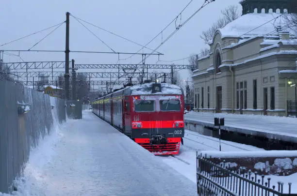 Red-orange electric train near the snow-covered platform next to the building of the old station gloomy winter morning