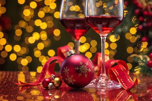 Red wine in wineglasses and Christmas ball against holiday lights background.