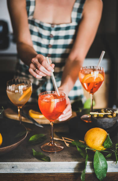 Young woman mixing ingredients in Spritz drink Young woman in checkered dress mixing ingredients in Spritz aperitif drink cocktail with orange in glass in kitchen. Summer refreshing drink concept aperitif photos stock pictures, royalty-free photos & images