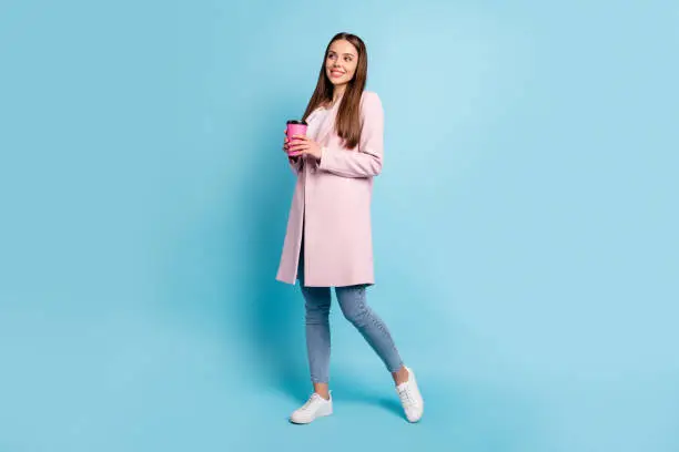 Photo of Full length photo of lovely dreamy girl having rest break holding take-out enjoy walk wearing topcoat denim jeans isolated over blue color background