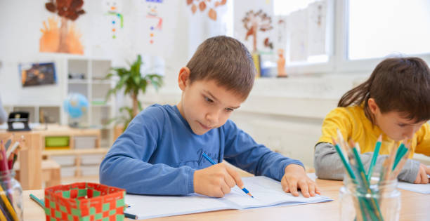 istock Boy drawing in his notebook sitting next to his classmate in bright classroom 1171896595