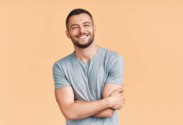 happy smiling handsome man with crossed arms looking to camera over beige background - sensuality horizontal indoors studio shot imagens e fotografias de stock