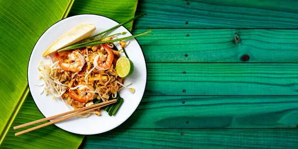 Pad Thai noodles with prawn are a world-famous delicacy, here, this colorful traditional dish is photographed directly above on a banana leaf on a textured wooden, colorful, vibrant turquoise background table.