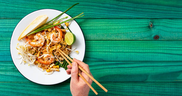 ターコイズ色の古い木製パネルテーブルの背景に箸でエビパッドタイ麺の世界的に有名なタイのレシピを食べる男。 - thailand thai culture thai cuisine pad thai ストックフォトと画像