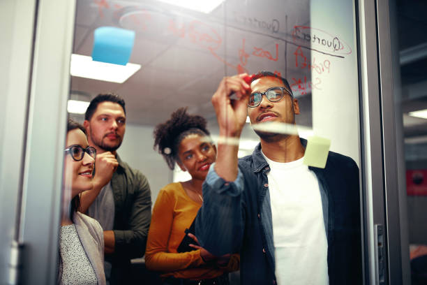 istock Multi ethnic happy creative business team looking at young man writing ideas on glass board with red marker at office 1171916384