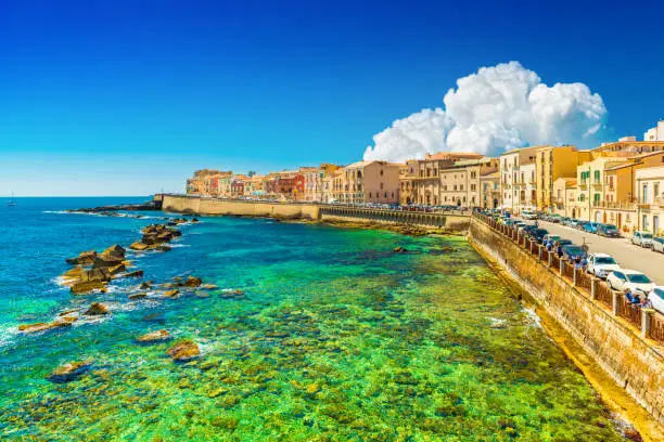 Beautiful cityscape of Ortygia (Ortigia), the historical center of Syracuse, Italy. Skyline of a European coastal town with turquoise transparent water and picturesque clouds in the sky.