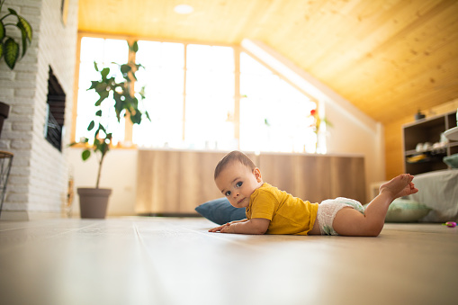 Adorable baby in diaper crawling on floor at apartment, looking at camera