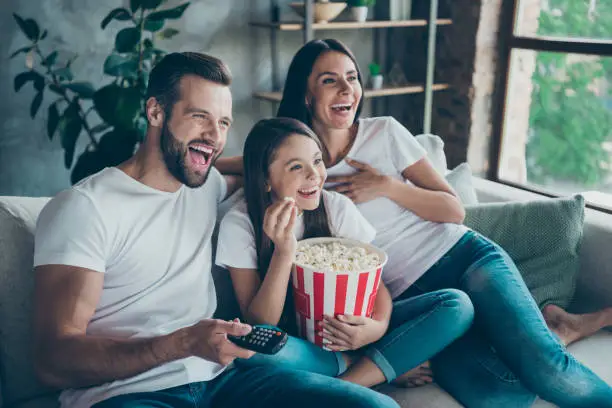 Photo of Portrait of nice attractive lovely positive glad cheerful cheery family wearing casual white t-shirts jeans denim sitting on sofa having fun watching funny video enjoying spending free time