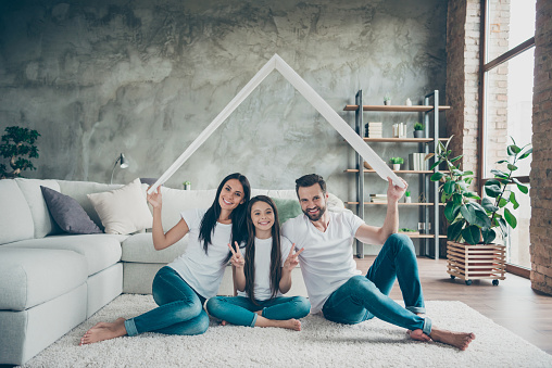 Portrait of nice attractive cheerful family in casual white t-shirts jeans sitting on carpet floor, holding in hand roof real estate ownership showing v-sign at industrial loft style interior living-room