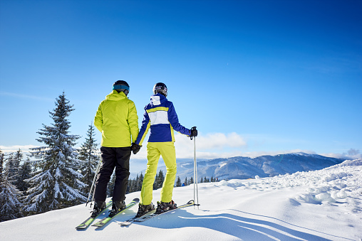 Couple of skiers holding hands on wooded mountain edge, enjoying wonderful beauty of nature at ski resort. Winter mountain panoramic landscape on background. Clear blue sky with copy space. Back view.