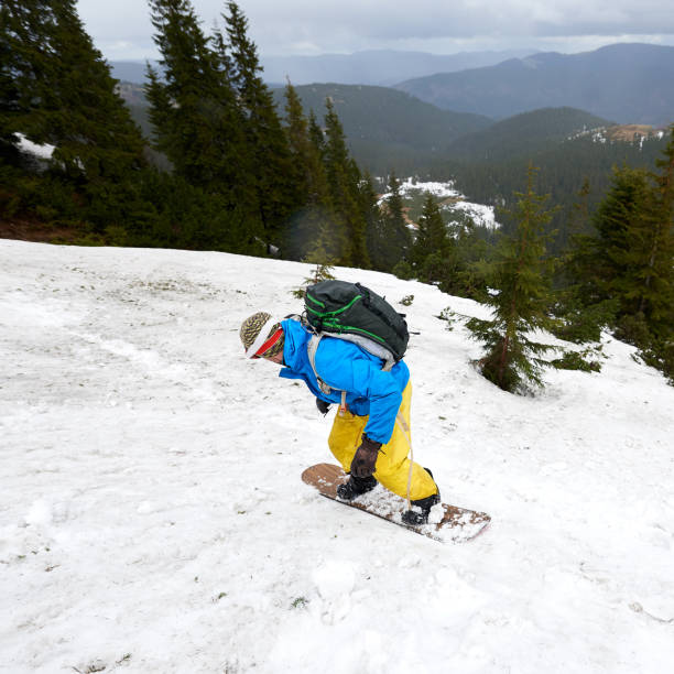 rückansicht des männlichen backpackers, der sich auf dem snowboard nach unten bewegt. bewaldete berge auf dem hintergrund. off-piste extremes snowboarden - back country skiing extreme skiing skiing ski stock-fotos und bilder