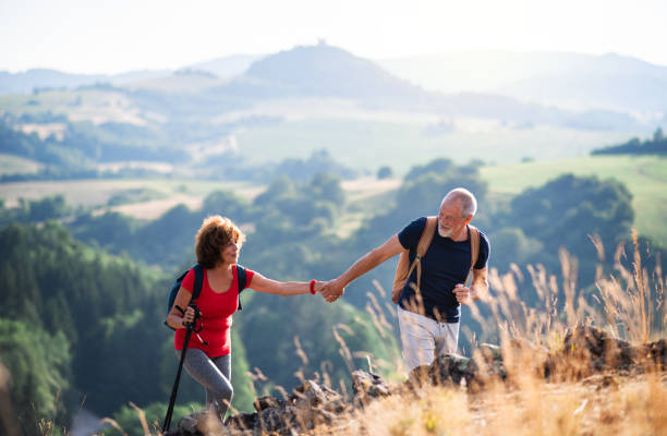 Senior tourist couple with backpacks hiking in nature, holding hands. A senior tourist couple with backpacks hiking in nature at sunset, holding hands. active lifestyle stock pictures, royalty-free photos & images