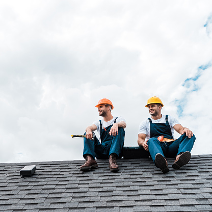cheerful handymen in helmets and uniform sitting on rooftop