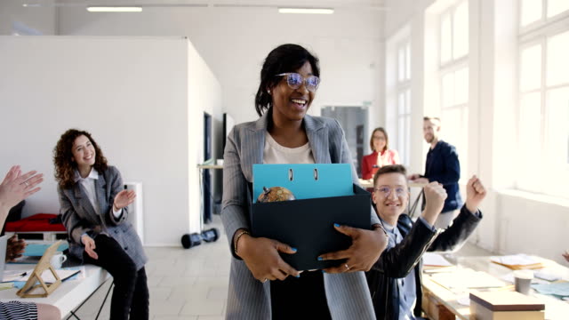 Beautiful young black female manager carrying office box, hired for a new job, multiethnic colleagues welcome and smile.
