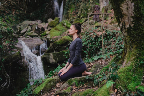 Young slim woman practicing yoga outdoors in moss forest on background of waterfall. Unity with nature concept. Girl meditates sitting Young slim woman practicing yoga outdoors in moss forest on background of waterfall. Unity with nature concept. Girl meditates sitting. sochi photos stock pictures, royalty-free photos & images