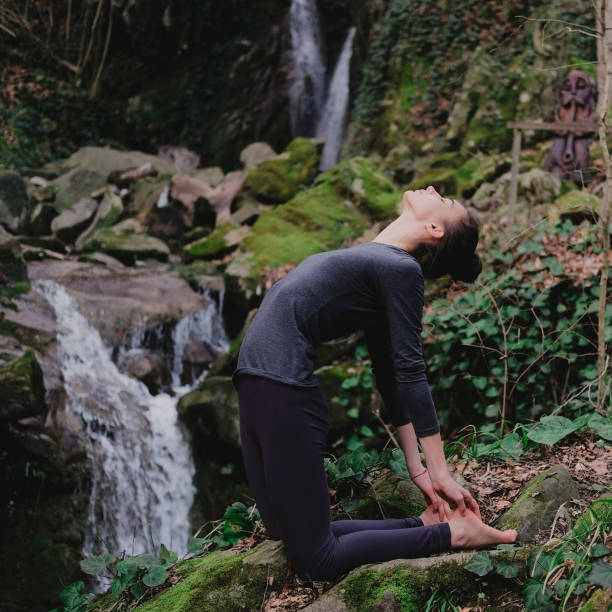 istock Young slim woman practicing yoga outdoors in moss forest on background of waterfall. Unity with nature concept. Girl in camel position 1171968921