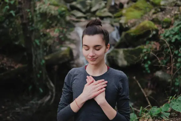 Young woman practicing breathing yoga pranayama outdoors in moss forest on background of waterfall. Unity with nature concept