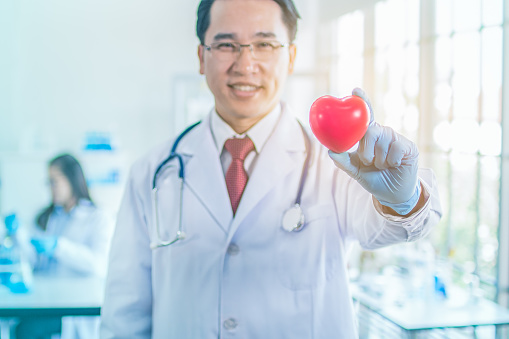 portarit smile face asia doctor man wearing coat uniform and stethoscope on neck holding red heart with blurred professional teamwork background in clinic hospital copy space.