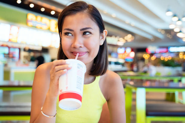 Asian woman having fast food, drinking with disposable cup in shopping mall food court stock photo