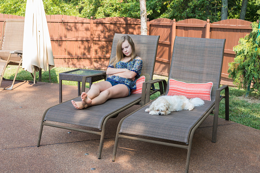 Blond teenage girl sits with her arms crossed in a chaise lounge chair next to her little white dog on a summer day, Indiana, USA