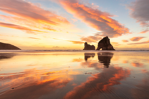 Winter sunset at Wharariki Beach at the top of New Zealand's South Island. The clouds are turned orange and red by the setting sun. The rocks are the Archway Islands. This beach is a popular tourist destination and is know for it's beautiful reflections of the islands.