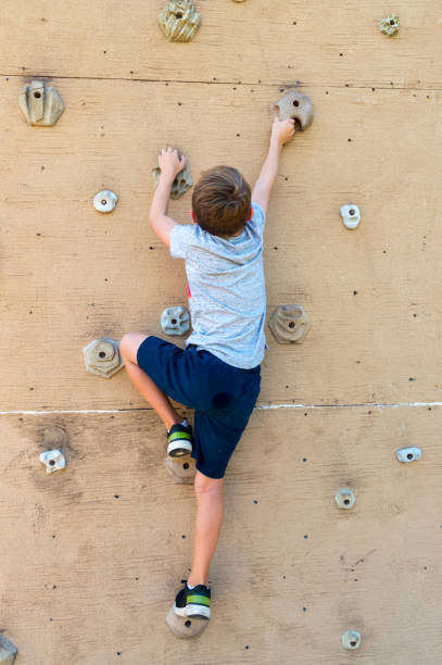 This photo shows a pre-teen boy climbing up a climbing wall in a park.  This was taken in the evening of a late summer day.