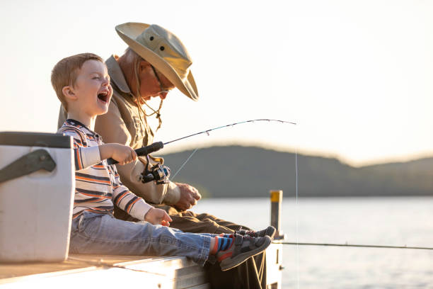 Grandfather and Grandson Fishing At Sunset in Summer A grandfather is teaching his grandson to fish during sunset in summer. They are both sitting on the dock and laughing. It is a beautiful summer day. Across the lake, there is a mountain. fishing rod photos stock pictures, royalty-free photos & images