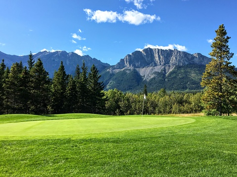 A view of a well manicured golf green with trees and the rocky mountains in the background.  It is a beautiful sunny day playing golf in Kananaskis.