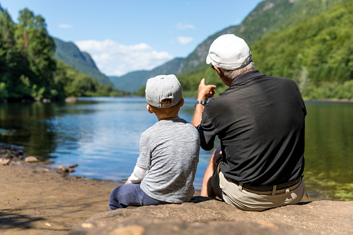 A grandfather and his grandson are sitting on a rock and contemplating the lake and mountains during their summertime vacations, Parc National de la Jacques-Cartier, Quebec, Canada