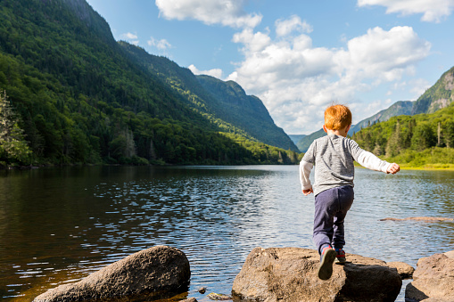 Little Redhead Boy Jumping on a Rock by the Lake and Mountains in Summer, Parc National de la Jacques-Cartier, Quebec, Canada
