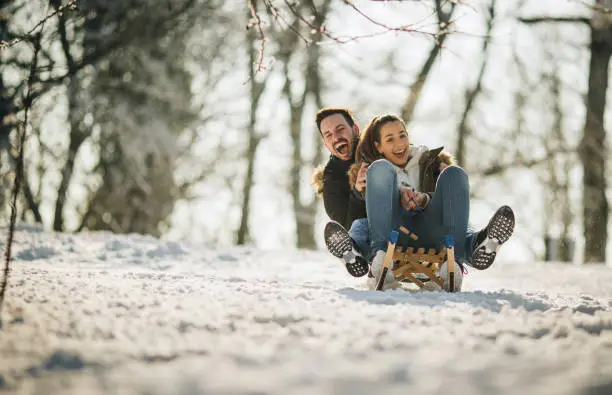 Young cheerful couple having fun on a sledge in winter day at the park. Copy space.