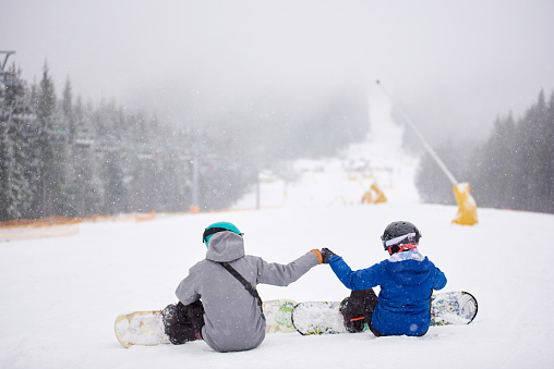 Couple snowboarders sitting on snow on ski run wooded slope at ski resort in snowfall with their boards on. Back view