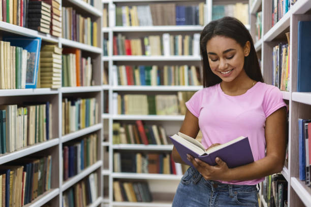 istock Smiling black girl reading book between bookshelves 1172141092