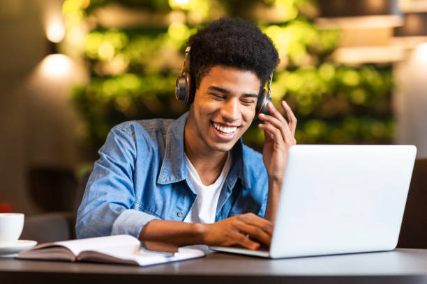 Cheerful teen guy with headset looking at laptop Cheerful black young guy with headset looking at laptop, having fun while studying, cafe interior music theory stock pictures, royalty-free photos & images