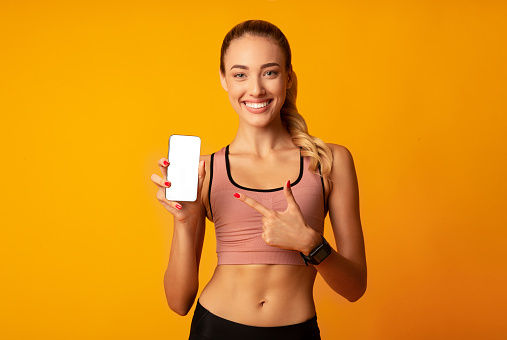 Sport Apps. Fitness Girl Pointing Finger At Cellphone Blank Screen On Yellow Background In Studio. Copy Space, Mockup