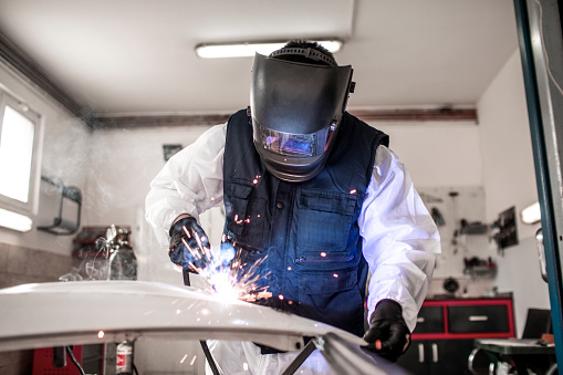 Car repairman using an arc welder to repair a damaged car part in a car body shop.